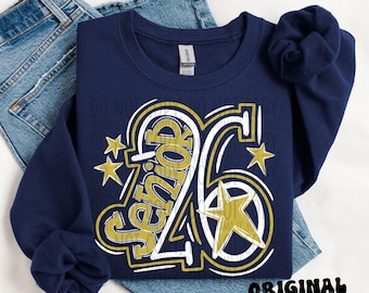Puede incluir: Sudadera azul marino con el texto "Senior 26" en dorado y blanco, junto con estrellas doradas. La sudadera está doblada sobre unos vaqueros azules. También se ven las palabras "Original Designer".