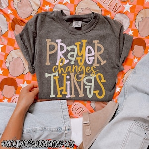 Może przedstawiać: Ciemnoszara koszulka z napisem "PRAYER changes THINGS" w kolorowej, zabawnej czcionce. Koszulka jest prezentowana na pomarańczowo-białym tle z wzorem. Widoczne są dłonie i dżinsy osoby. Odzież codzienna.