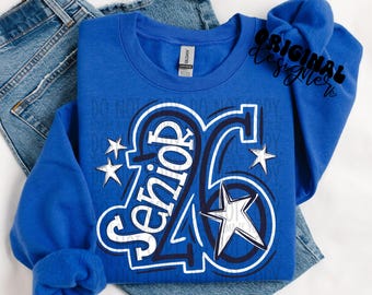 Puede incluir: Una sudadera azul real con el texto "Senior '26" en un diseño blanco y azul marino, junto con gráficos de estrellas. La sudadera está doblada sobre un par de vaqueros azules. Las palabras "Original Designer" están escritas en negro.