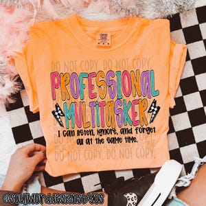 Puede incluir: Camiseta naranja con el texto "PROFESSIONAL MULTITASKER" en letras de colores. El texto está rodeado de un patrón de cuadros blanco y negro y un rayo. El texto de abajo dice "Puedo escuchar, ignorar y olvidar todo al mismo tiempo."