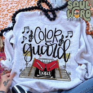 May include: A light grey sweatshirt featuring the words "Color Guard" in black script, accompanied by musical notes and a band graphic. The band graphic includes red flags and the word "band".