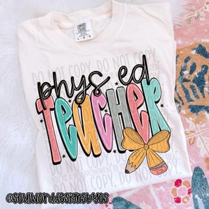Può includere: T-shirt color crema con le parole "phys ed teacher" in lettere a bolle colorate. Una matita gialla e una grafica a fiocco sono sotto il testo. La maglietta ha un'etichetta Comfort Colors.