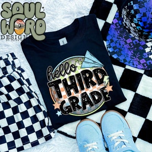 Puede incluir: Camiseta negra con un gráfico de estilo retro que dice "hello third grade" con un patrón a cuadros y un avión de papel.