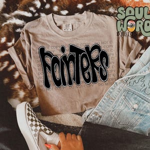 May include: A brown t-shirt with the word "Fointers" printed in black on the front. The shirt is on a fuzzy brown surface with a pair of jeans and a pair of checkered Vans shoes.