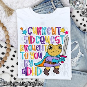 May include: White t-shirt with the text "CURRENT SIDEQUEST BROUGHT TO YOU BY ADHD" in colorful letters. A cartoon frog wearing a cape and holding a sword is featured in the design. The shirt is displayed on a pair of jeans.