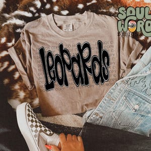 May include: A brown t-shirt with the word "Leopards" printed in black and white graffiti style lettering. The shirt is laid out on a faux fur surface with a pair of checkered Vans shoes and a denim jacket.