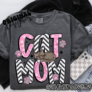 Puede incluir: Una camiseta gris con un dise&ntilde;o rosa y negro que dice "Cat Mom" con un gr&aacute;fico de un gato durmiendo. El dise&ntilde;o presenta un patr&oacute;n de chevron y flores rosas.