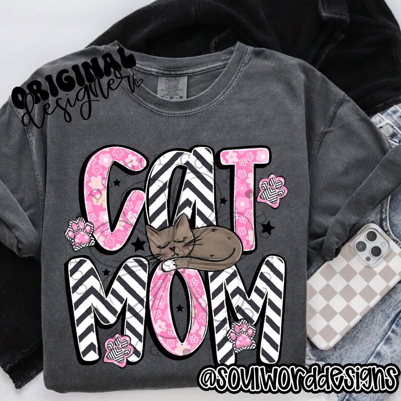 Puede incluir: Una camiseta gris con un dise&ntilde;o rosa y negro que dice "Cat Mom" con un gr&aacute;fico de un gato durmiendo. El dise&ntilde;o presenta un patr&oacute;n de chevron y flores rosas.