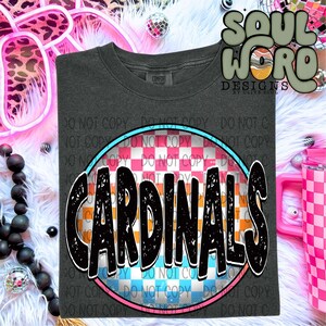 Puede incluir: Una camiseta gris oscuro con un diseño circular que presenta un patrón a cuadros en rosa, azul y blanco. La palabra "CARDINALS" está escrita en negro con un aspecto desgastado sobre el patrón a cuadros.