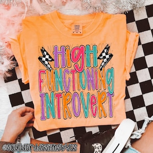 Könnte beinhalten: Orangefarbenes T-Shirt mit dem Text "High Functioning Introvert" in bunten Buchstaben mit Blitzen auf beiden Seiten.