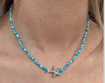 Peut inclure: Un collier de perles avec un pendentif en forme d'étoile de mer argenté. Le collier est composé de petites perles colorées dans les tons de bleu, rose et vert. Le collier est porté autour du cou.