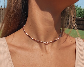May include: A beaded choker necklace with a mix of white, brown, and dark brown beads. The necklace is worn around the neck and is a delicate piece of jewelry. The beads are small and evenly spaced.