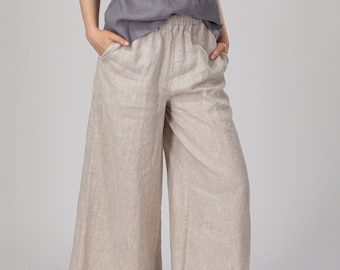 May include: A woman is wearing a gray linen tank top and beige linen wide-leg pants. She is standing with her hands in her pockets and looking at the camera.