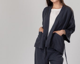 Può includere: Un completo pantalone in lino blu scuro con una giacca a maniche lunghe e pantaloni larghi. La giacca ha una chiusura a cravatta e tasche. I pantaloni hanno una chiusura a cravatta in vita. La modella indossa sandali dorati.