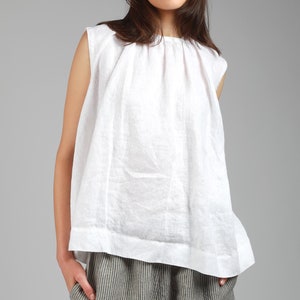 May include: A white linen sleeveless top with a gathered neckline and a relaxed fit. The top has a slightly rounded hemline and is shown on a model wearing it with striped trousers.