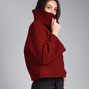 Puede incluir: Una chaqueta roja corta con cuello alto y mangas largas. La chaqueta está hecha de una tela suave y texturizada.