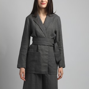 May include: A dark grey linen suit featuring a wrap-around jacket with a matching belt and wide-leg trousers. The jacket has long sleeves and pockets. The outfit is made of a soft, textured fabric, and the model is wearing neutral-toned sandals.