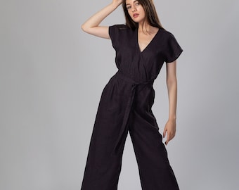 May include: A woman wearing a dark gray linen jumpsuit with a wrap-style bodice and a wide belt. She is wearing brown sandals and has her hair pulled back.