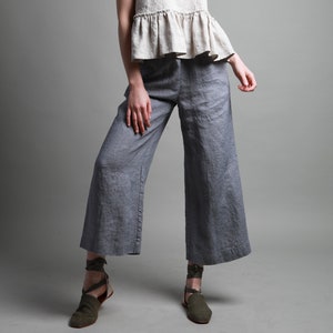 May include: A woman wearing a light beige linen top with a ruffle hem and wide-legged gray linen pants. She is wearing green ankle strap sandals.