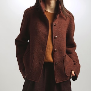 Puede incluir: Chaqueta de lana marrón con cuello alto, cierre de botones y dos bolsillos delanteros. La chaqueta se lleva sobre un top naranja y se combina con una falda marrón oscuro. La chaqueta tiene un diseño clásico y entallado.