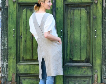 May include: A woman wearing a light brown linen apron with a large pocket on the front, standing in front of a green wooden door. The apron has a criss-cross strap design at the back.