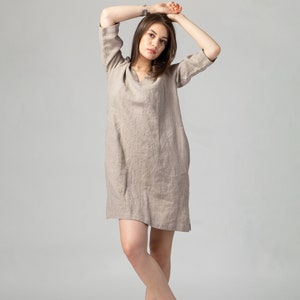 May include: A woman wearing a beige linen dress with a v-neck and 3/4 sleeves. She is standing with her arms crossed and is wearing brown sandals.