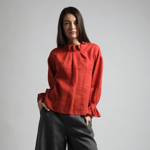 May include: A red long-sleeved blouse with a ruffled collar and cuffs. The blouse is paired with dark gray wide-leg pants. The outfit is made of a soft, flowing fabric, perfect for a casual or semi-formal look.
