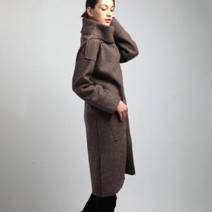 Puede incluir: Un abrigo largo de lana marr&oacute;n con un cuello alto y ancho y grandes bolsillos delanteros. El abrigo llega a la rodilla y tiene mangas largas. La modelo lleva pantalones y botas negras. El abrigo es de color liso y parece estar hecho de una tela suave y texturizada.