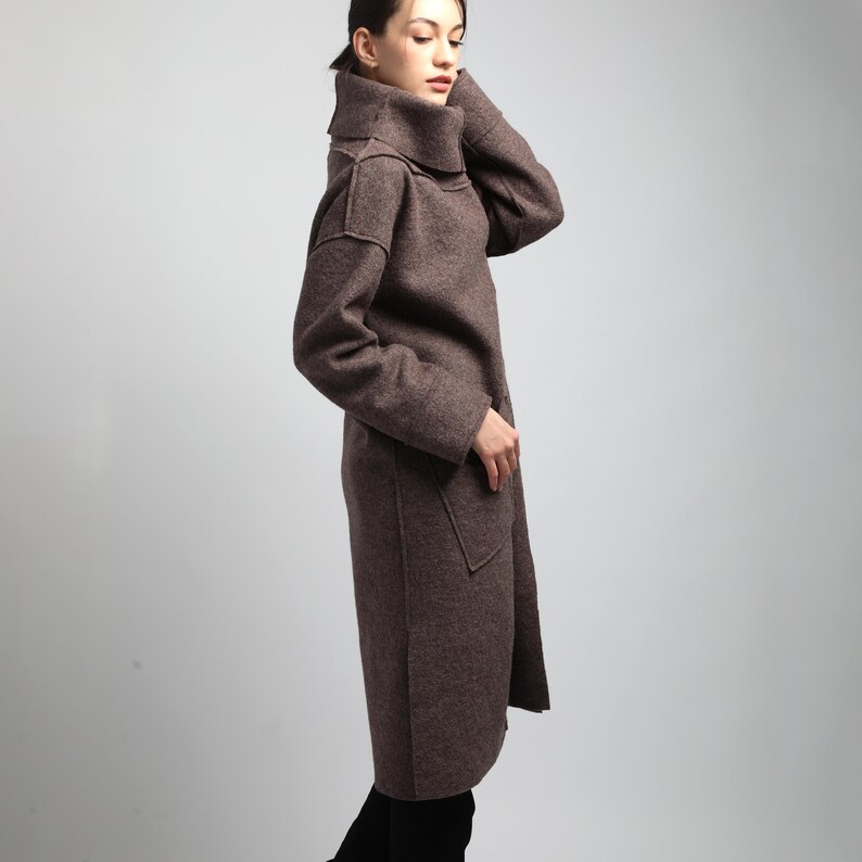 Puede incluir: Un abrigo largo de lana marr&oacute;n con un cuello alto y ancho y grandes bolsillos delanteros. El abrigo llega a la rodilla y tiene mangas largas. La modelo lleva pantalones y botas negras. El abrigo es de color liso y parece estar hecho de una tela suave y texturizada.
