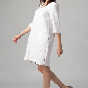 May include: A white linen dress with a v-neck and 3/4 sleeves. The dress has a side slit and is worn with brown espadrille sandals.
