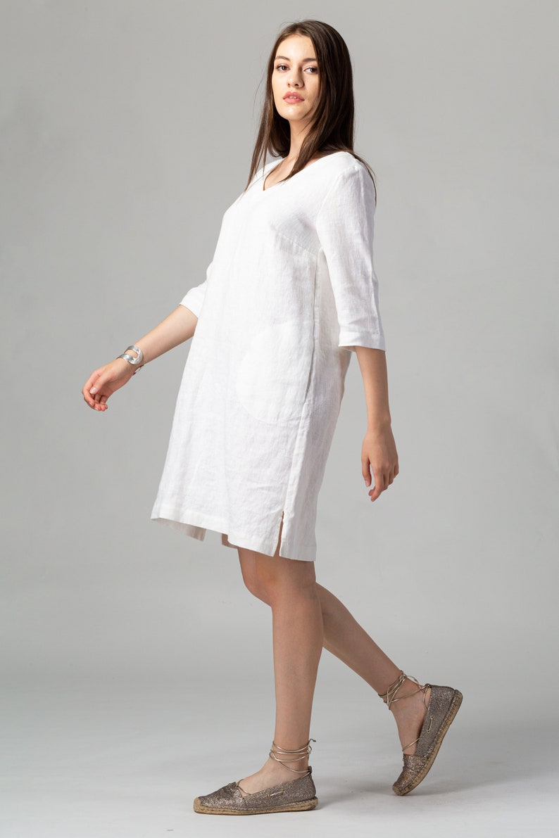 May include: A white linen dress with a v-neck and 3/4 sleeves. The dress has a side slit and is worn with brown espadrille sandals.