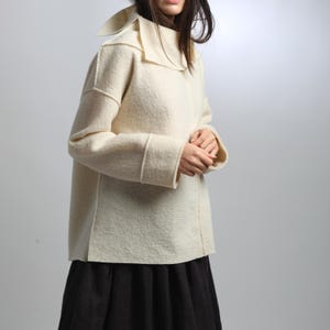 May include: A cream-colored, wool-like jacket with a wide collar and long sleeves. The jacket has a boxy shape and is paired with a black skirt. The sleeves have a cuff detail. The jacket is a neutral color and appears to be warm.