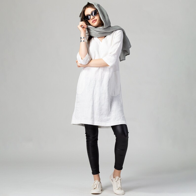 May include: A woman wearing a white linen tunic dress with a grey scarf and black leggings. She is wearing sunglasses and white trainers.