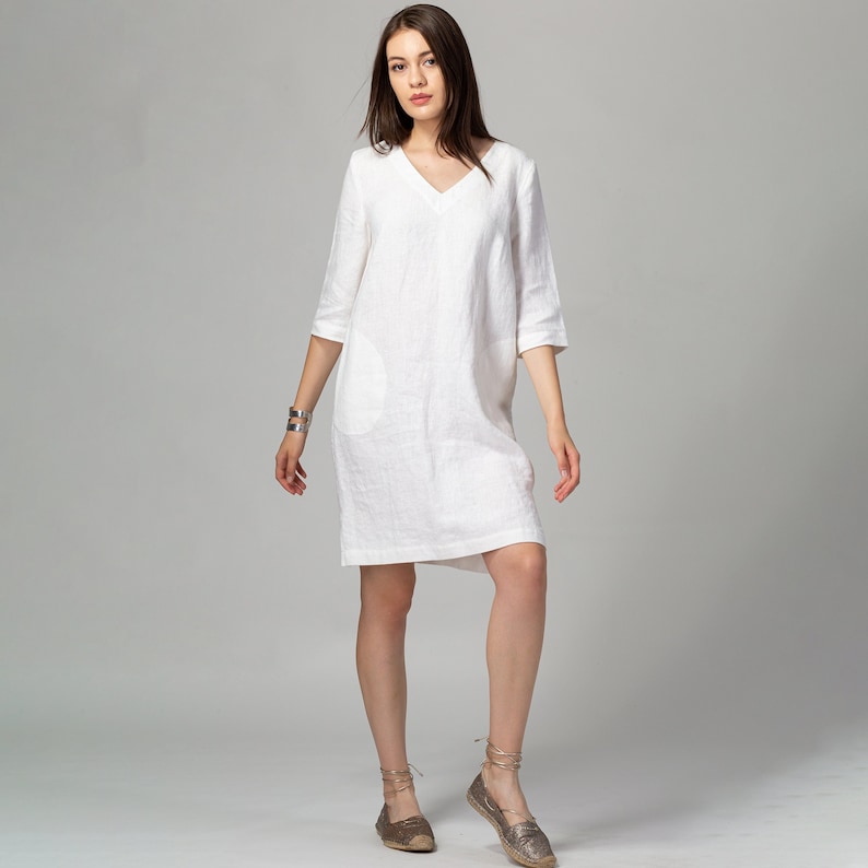 May include: A white linen dress with a v-neck and short sleeves. The dress has a pocket on the left side and a relaxed fit.