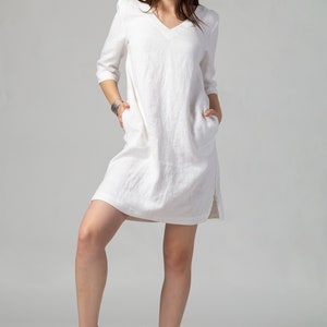 May include: A white linen dress with a v-neckline and 3/4 sleeves. The dress has pockets on both sides and a relaxed fit. The model is wearing the dress with gold sandals.