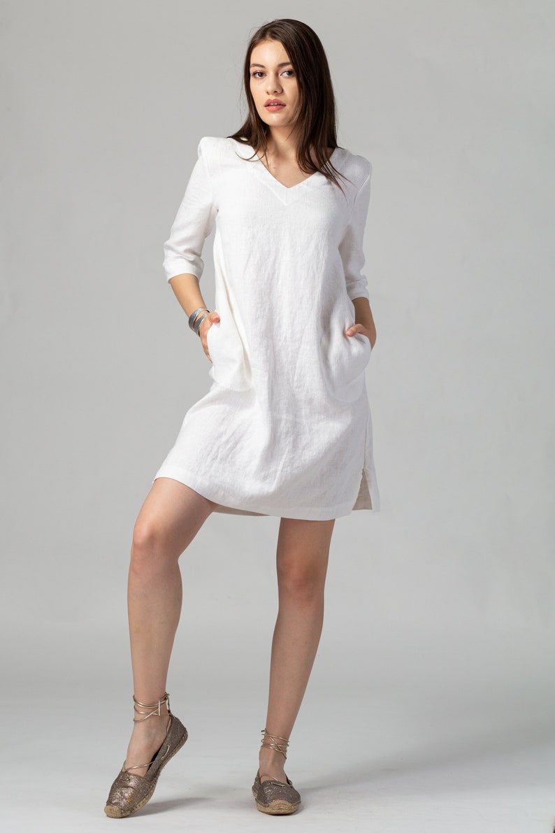 May include: A white linen dress with a v-neckline and 3/4 sleeves. The dress has pockets on both sides and a relaxed fit. The model is wearing the dress with gold sandals.