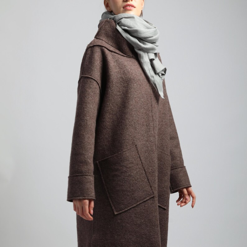 Puede incluir: Un abrigo de lana marr&oacute;n con grandes bolsillos delanteros y cuello alto. Una bufanda gris claro est&aacute; drapeada alrededor del cuello. El abrigo tiene un corte holgado y mangas largas.