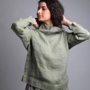 Pode incluir: Um suéter de linho verde com gola alta e mangas compridas. O suéter é solto e tem um estilo relaxado e casual.