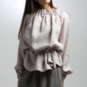 May include: A light mauve linen blouse with long sleeves and a gathered neckline. The blouse features a tie-waist design and elastic cuffs. The sleeves are long and voluminous, and the fabric has a soft, textured appearance. The blouse is paired with a gray skirt.