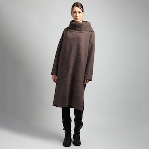 May include: A long, brown wool dress with a high, cowl neck and long sleeves. The dress has two large front pockets and a knee-length hem. The model is wearing black pants and black boots. The dress is a neutral color and appears to be warm and comfortable.