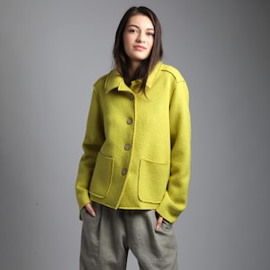 Puede incluir: Chaqueta de color chartreuse con botones, dos bolsillos delanteros y cuello. La chaqueta es de tejido texturizado y se combina con pantalones grises. La modelo posa frente a un fondo gris.