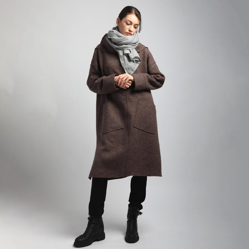 Puede incluir: Un abrigo largo marr&oacute;n con capucha y dos bolsillos delanteros. El abrigo se lleva con una bufanda gris, pantalones negros y botas negras. El abrigo parece estar hecho de un material grueso y c&aacute;lido, adecuado para el clima fr&iacute;o.