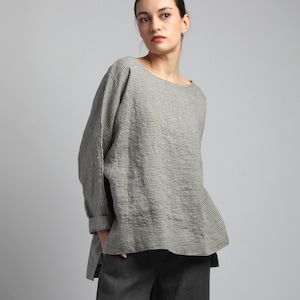 Peut inclure: Un haut à manches longues avec un motif de fines rayures horizontales dans des tons de gris et de blanc. Le vêtement présente une large encolure arrondie, des épaules tombantes et un ourlet asymétrique avec des fentes latérales, porté avec un pantalon gris foncé.