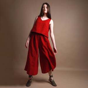 Puede incluir: Una mujer lleva un mono de lino rojo de pierna ancha. El mono tiene un escote en V y una parte superior recortada. La mujer est&aacute; de pie sobre un fondo marr&oacute;n claro.
