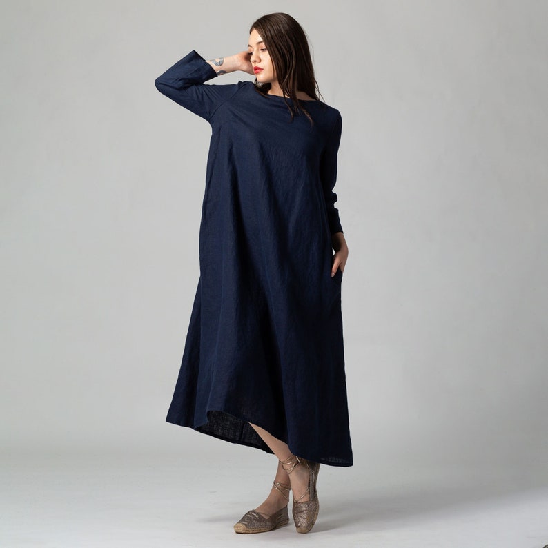 May include: A long, navy blue, linen dress with long sleeves and a loose fit. The dress has a slightly asymmetrical hemline and is worn with brown sandals.