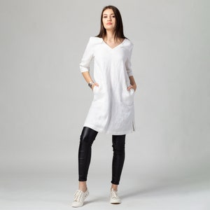 May include: A white linen tunic dress with a v-neckline and pockets. The dress is worn with black leggings and white trainers.