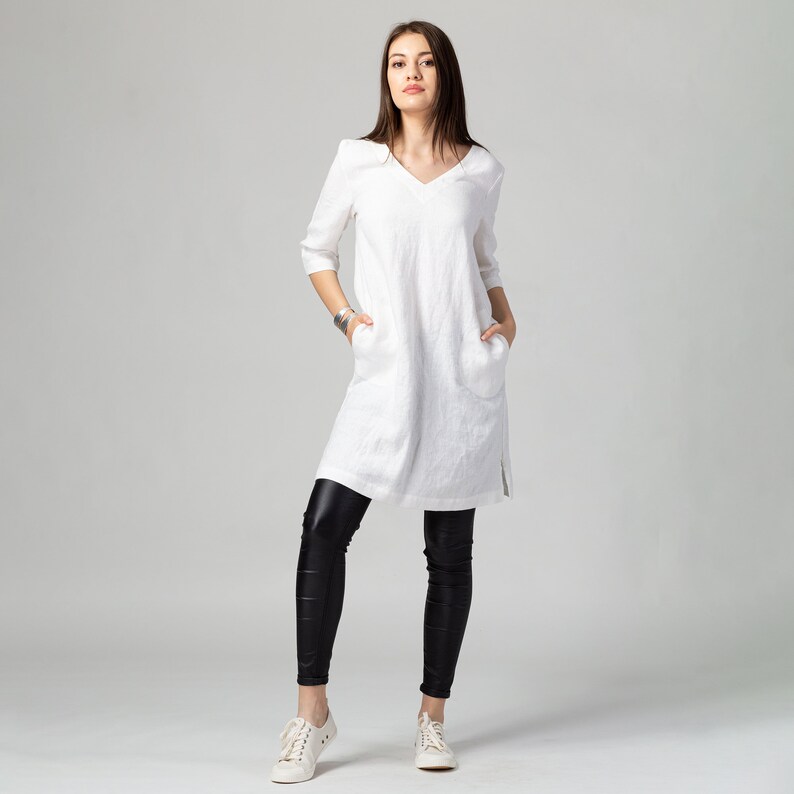 May include: A white linen tunic dress with a v-neckline and pockets. The dress is worn with black leggings and white trainers.
