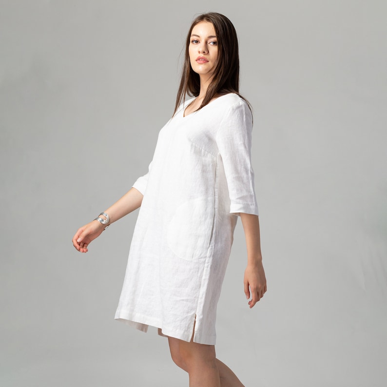 May include: A white linen dress with a v-neck and 3/4 sleeves. The dress has a side pocket and a side slit.