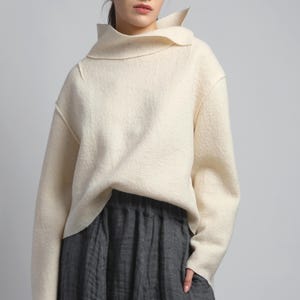 May include: Cream-colored sweater with a unique, folded collar and long sleeves. The sweater has a textured appearance and a relaxed fit. Paired with a dark gray, pleated skirt. The outfit is simple and elegant, suitable for various occasions.