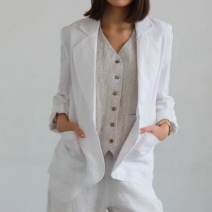 Peut inclure: Un blazer blanc en lin avec une fermeture simple et deux poches sur le devant. Le blazer est porté sur un gilet rayé.
